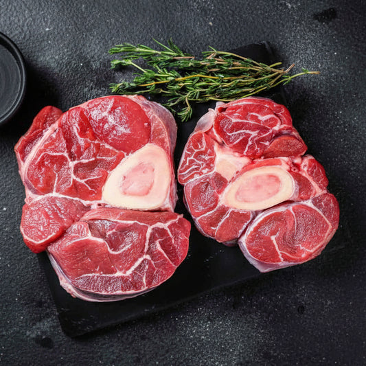 Two raw beef shanks on a dark surface with herbs and red pepper flakes.
